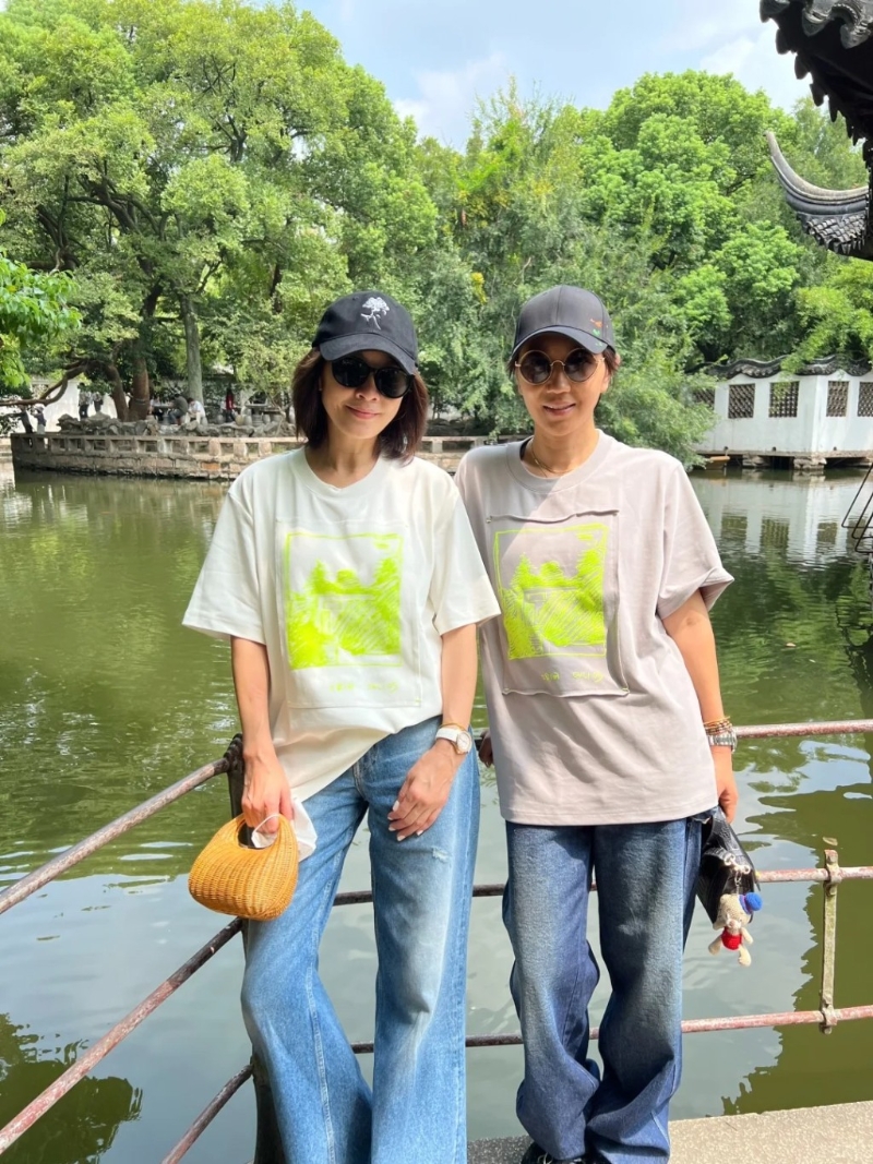劉嘉玲頭戴黑色cap帽，身穿一件印有螢光圖案的白色短袖T恤、淺藍色牛仔長褲，身上拎住一個編織包包，非常休閑地在園林影靚相。